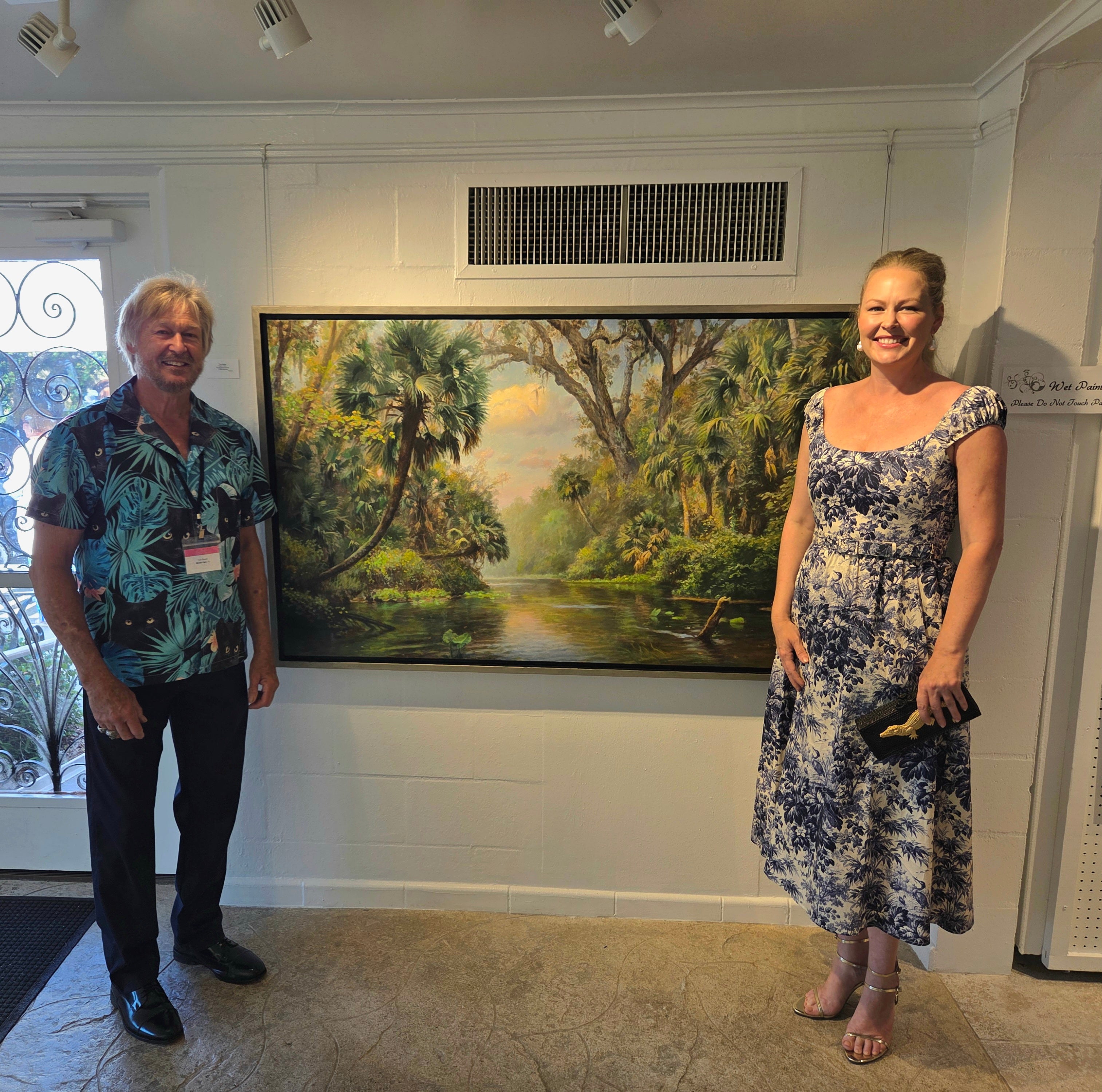 Two people standing in front of a large painting of a landscape.